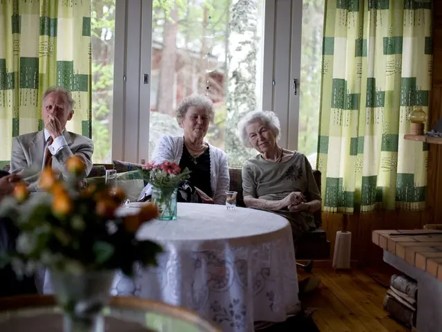 There are group of old people sitting on sofa and a table in front of them with a flower vase on...