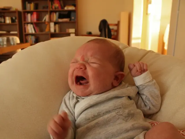 In this image we can see a baby crying and lying on a sofa. In the back there are cupboards. Inside...