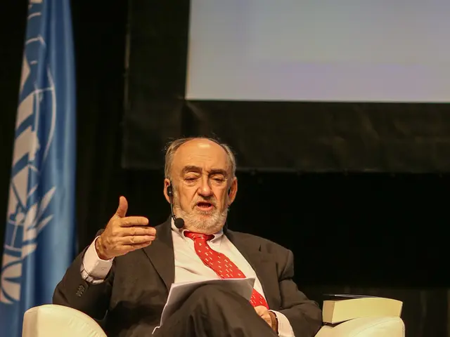 In this image I can see a man wearing suit, sitting on the chair and holding a paper in his hand...